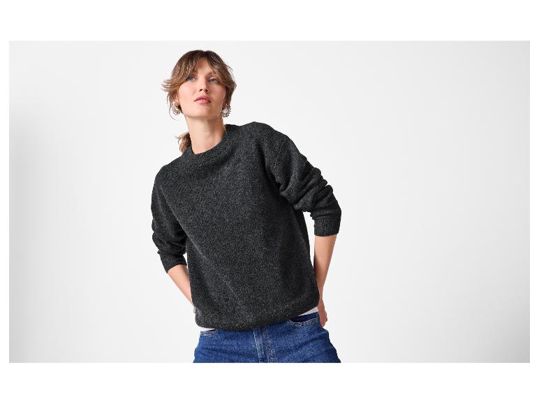 Vrouw in een donkergrijze trui en blauwe jeans, omhoogkijkend.