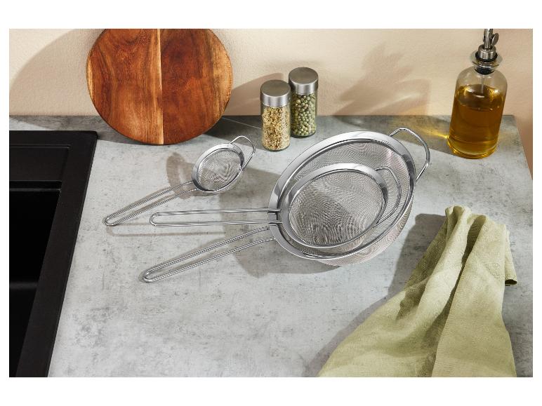 Set van drie roestvrijstalen zeven in verschillende maten op een aanrecht met snijplank en kruiden.