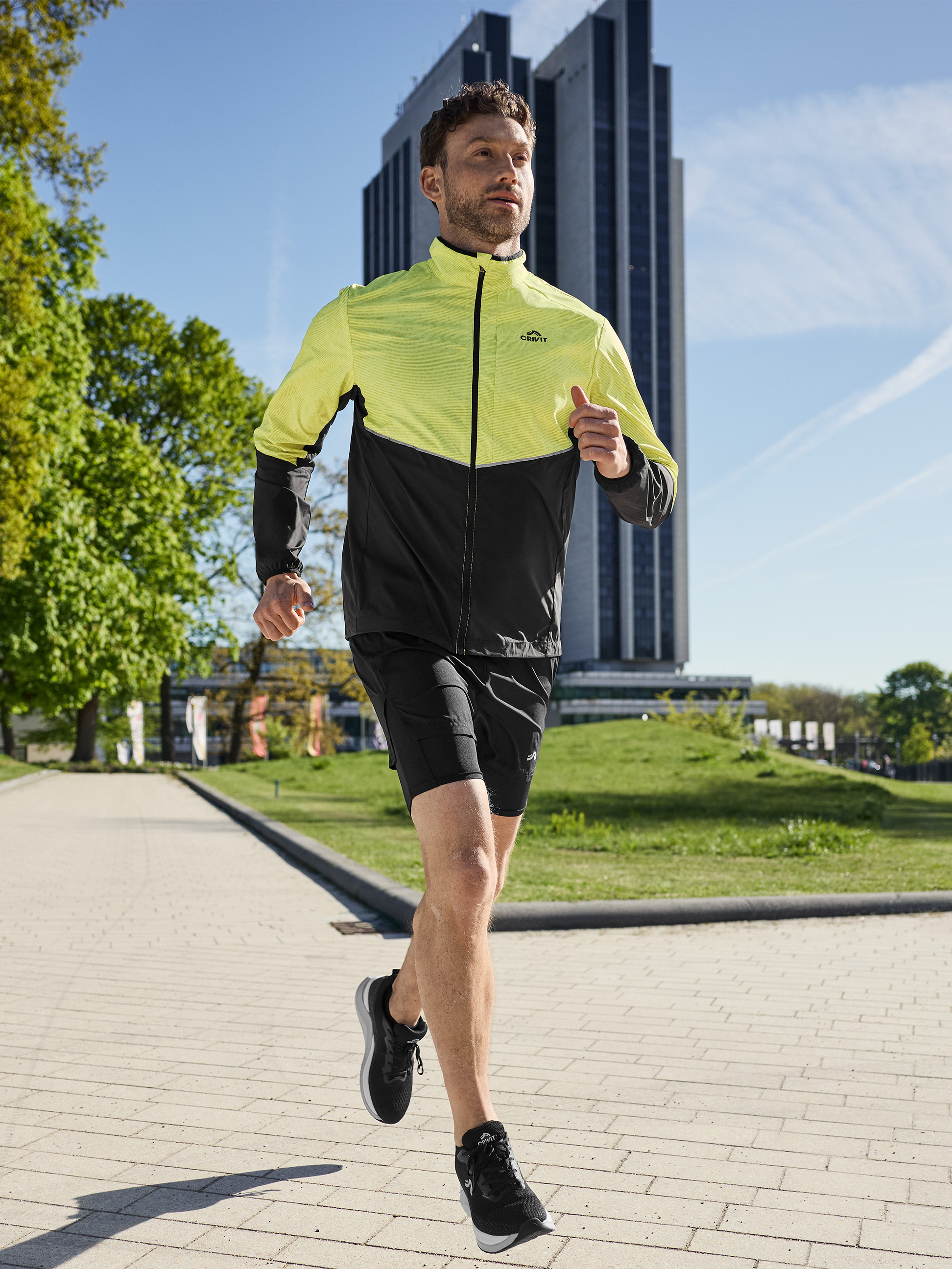 Man in een geel-zwart hardloopjack en zwarte shorts, buiten aan het hardlopen.