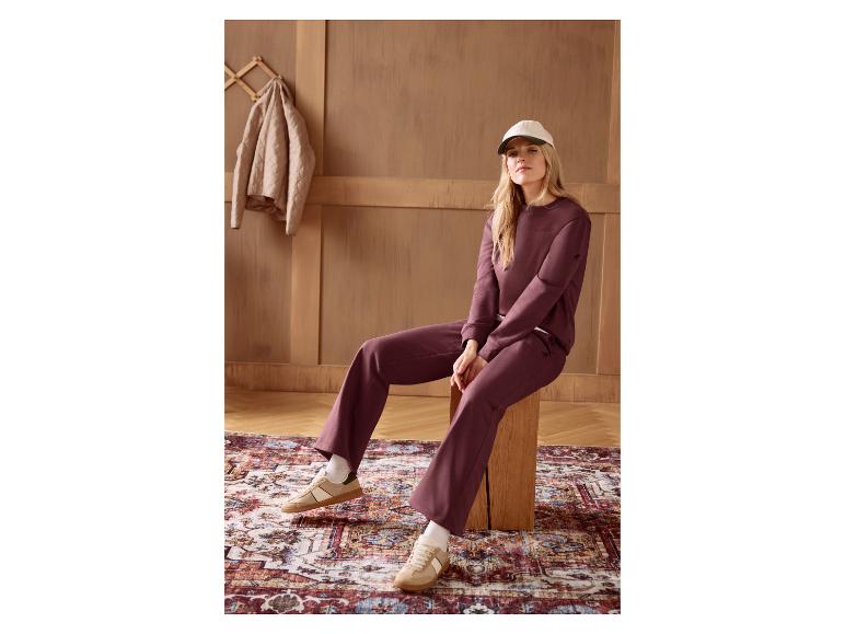 Vrouw in bordeauxrode sweater en bijpassende broek, beige sneakers en pet, zittend op een houten kruk.