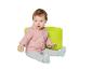 Baby in roze hoodie en grijze broek, zittend op een groene poef met een houten blokje.