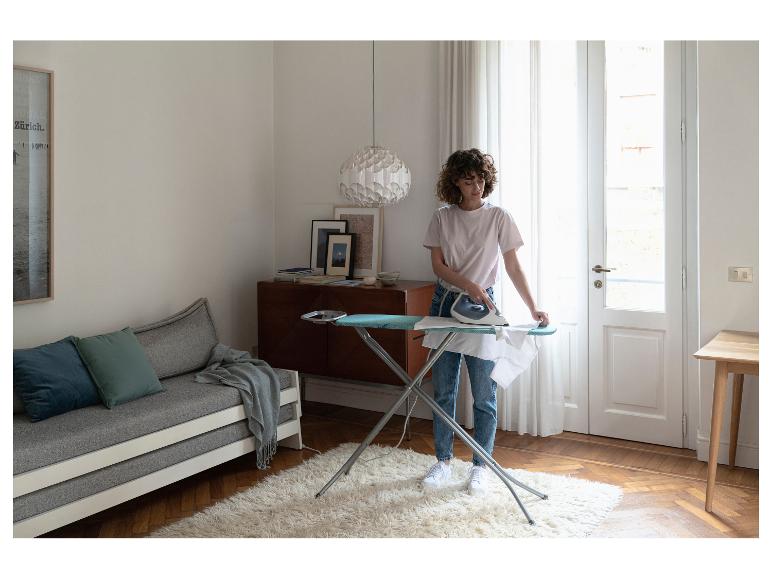 Vrouw strijkt wit overhemd op strijkplank in woonkamer met bank en vloerkleed.