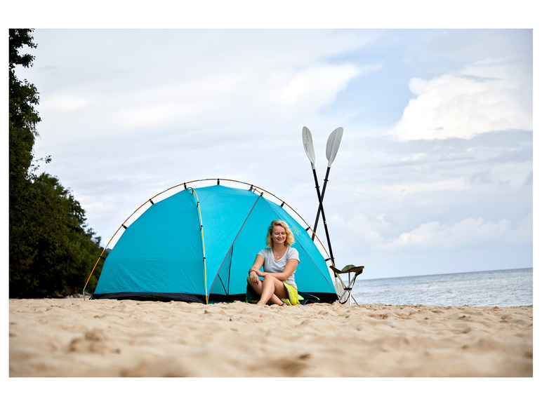 Een vrouw zit voor een blauwe tent op het strand met twee peddels ernaast.