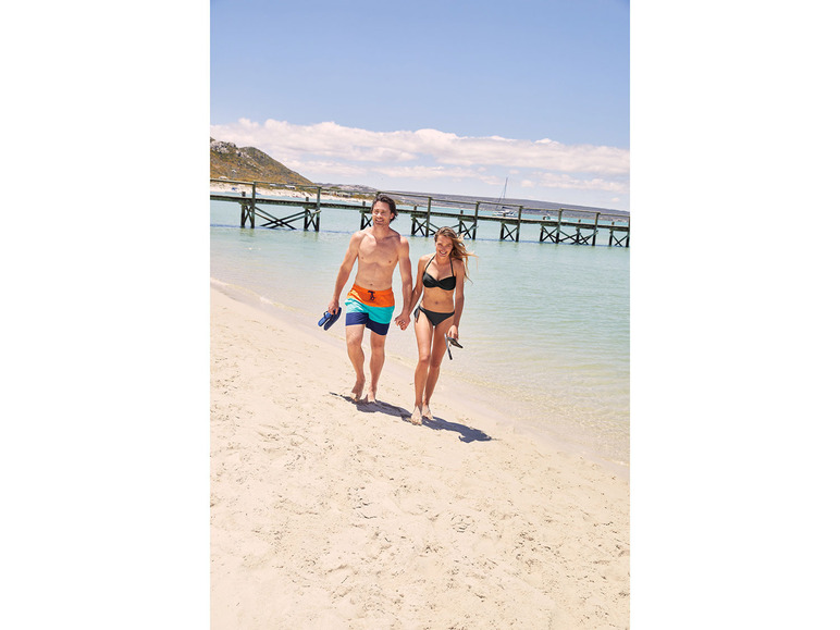 Een stel in zwemkleding loopt hand in hand over het strand.