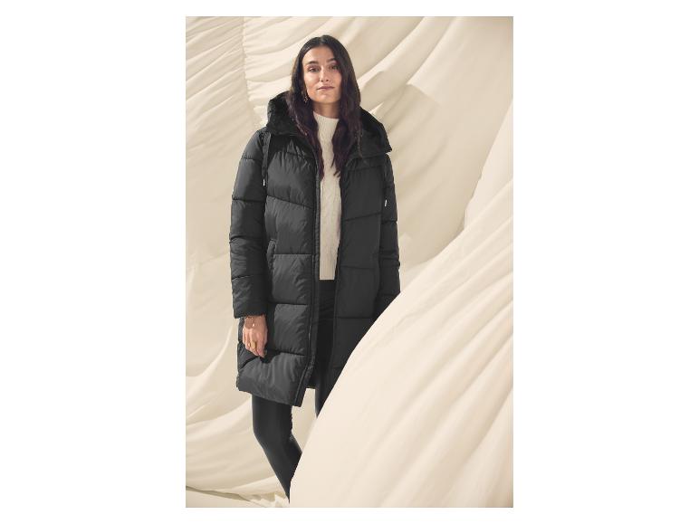 Vrouw in een lange zwarte jas met een witte trui, op een beige achtergrond.