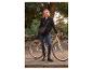 Vrouw in zwarte jas en jeans, staand naast een fiets met een schoudertas.