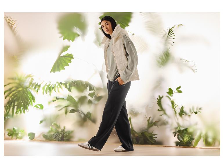 Vrouw in beige bodywarmer, grijze sweater en zwarte broek met planten op de achtergrond.