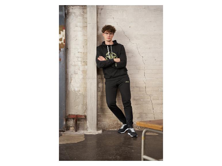 Man in zwarte Jeep hoodie en joggingbroek leunend tegen een muur.