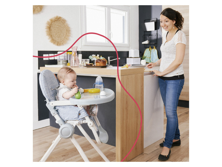 Een baby zit in een blauwe kinderstoel en eet, terwijl een vrouw in de keuken staat.