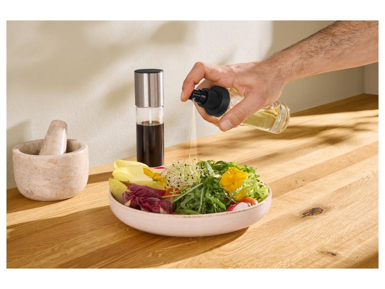 Een hand spuit olie op een saladekom, met een vijzel en stamper en een fles azijn op de achtergrond.