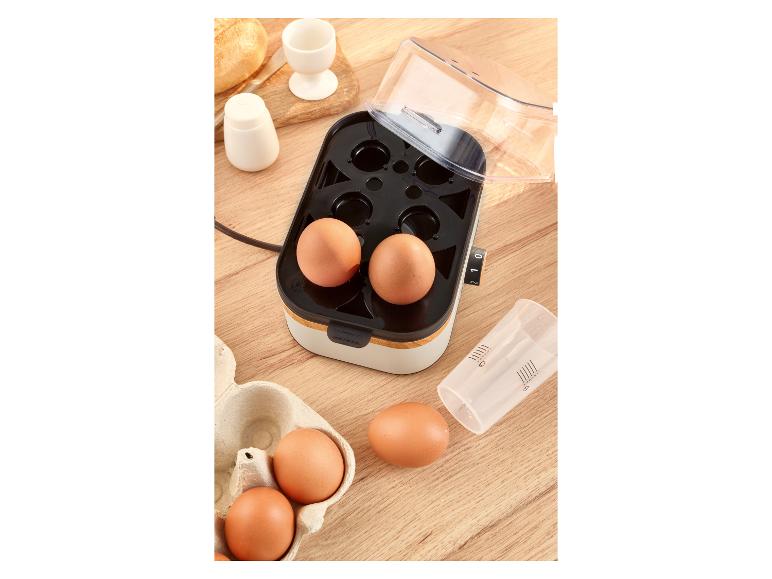 Eierkoker met eieren, maatbeker, zoutvaatje, eierdopje en brood op een houten tafel.