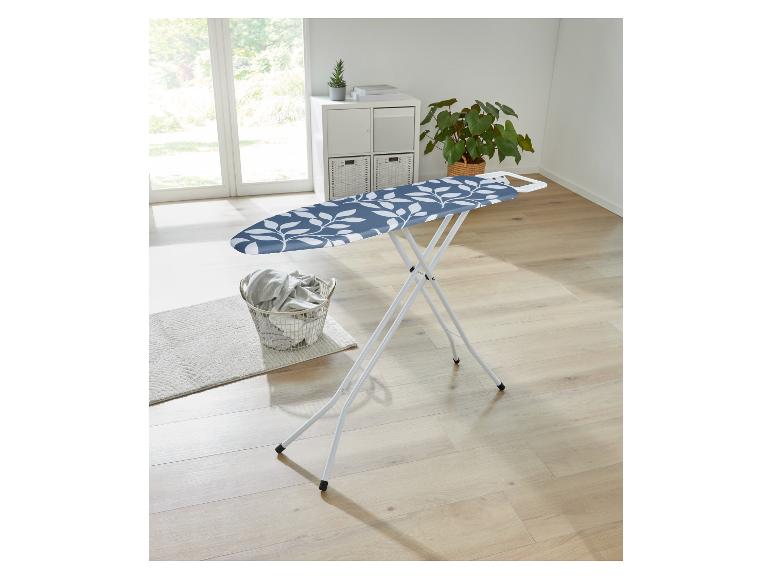 Strijkplank met blauw wit-blad motief, wasmand en planten in een lichte kamer.