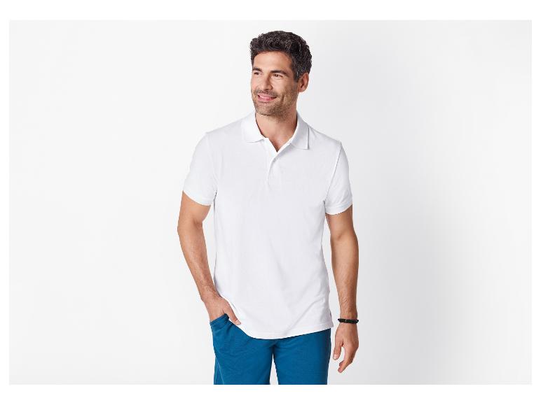 Man in een wit poloshirt en blauwe korte broek, lachend, met zijn hand in zijn zak.
