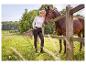 Vrouw in wit shirt en donkere broek met paard bij houten hek in landelijk landschap.