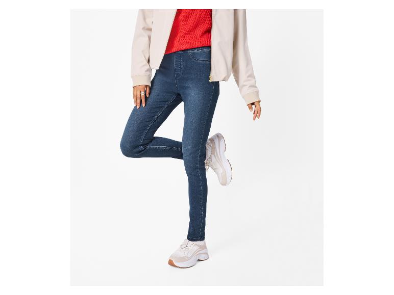Vrouw in beige jasje, rode trui en blauwe skinny jeans met beige sneakers.