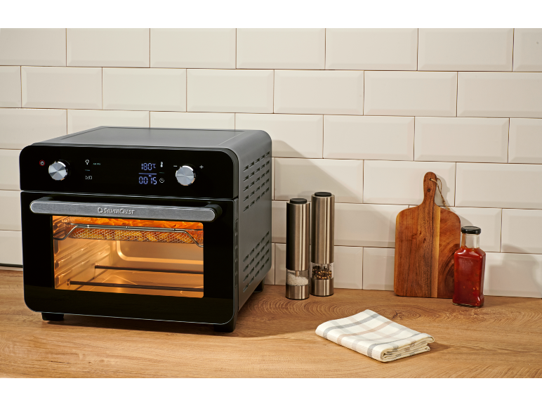 Zwarte airfryer op een houten aanrecht met witte tegels, zout en peper, snijplank en saus.