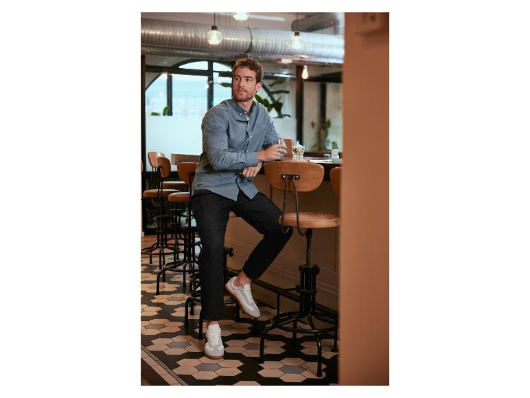 Een man in een lichtblauw overhemd, zwarte broek en witte sneakers zit aan een bar.