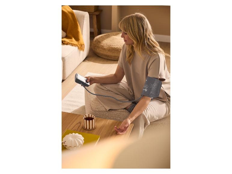 Vrouw meet bloeddruk met bovenarmbloeddrukmeter, zittend op de grond met een kopje thee.