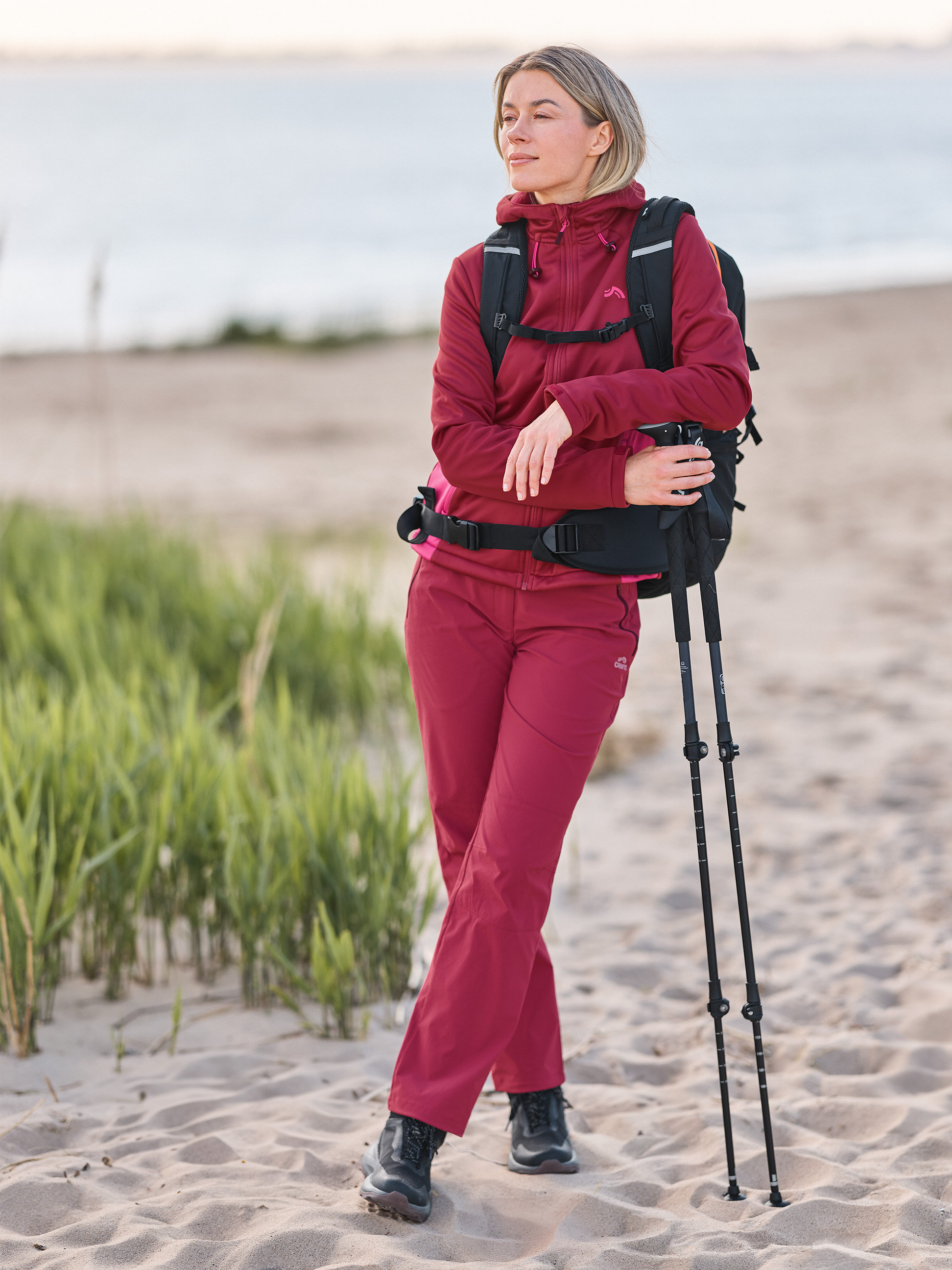 Vrouw in wandelkleding en rugzak op het strand, met wandelstokken.