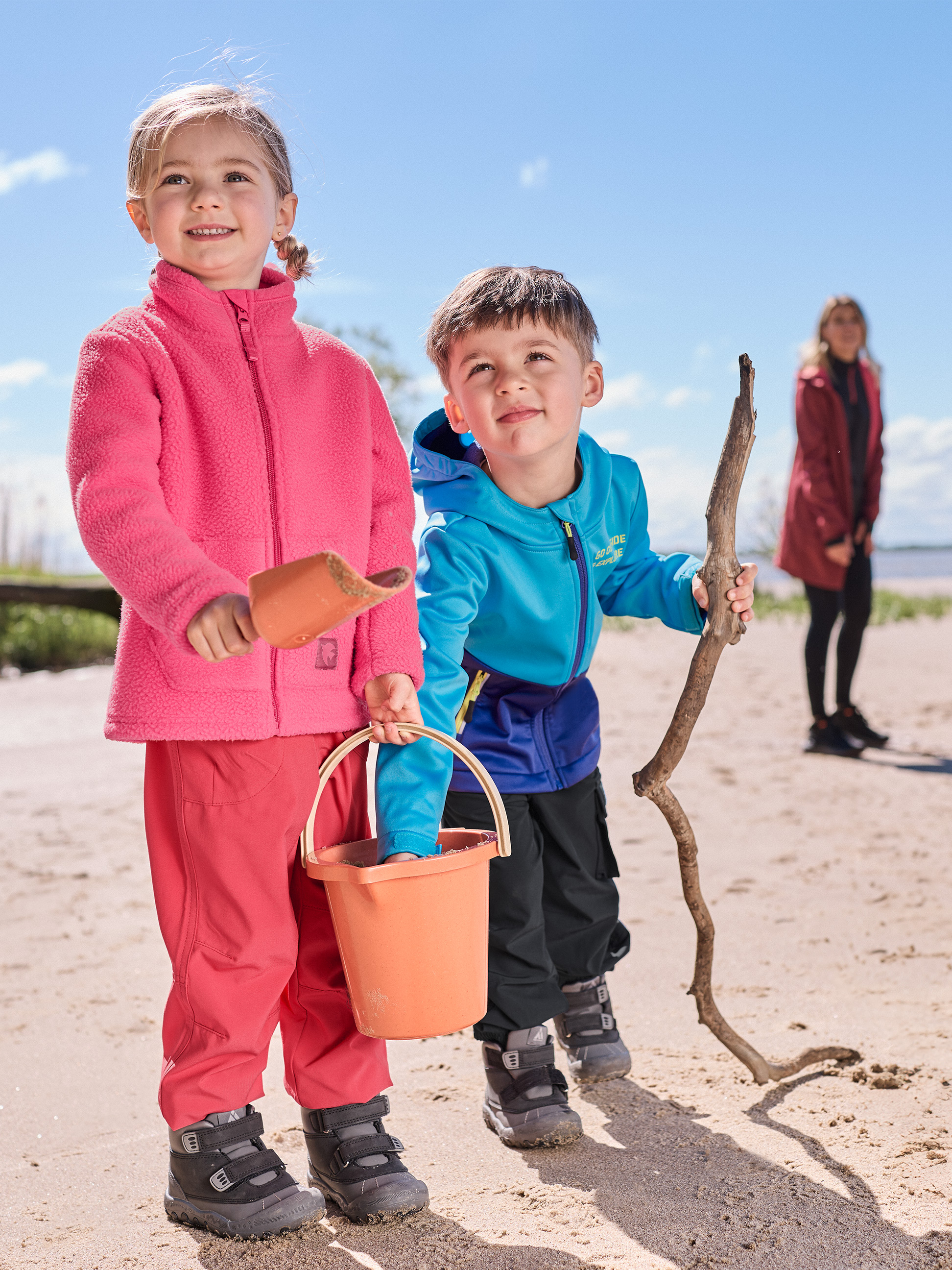 Twee kinderen spelen op het strand, gekleed in roze en blauwe fleeces en waterdichte broeken.
