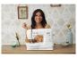 Vrouw lacht achter een naaimachine, omringd door decoratieve items en naaiaccessoires.