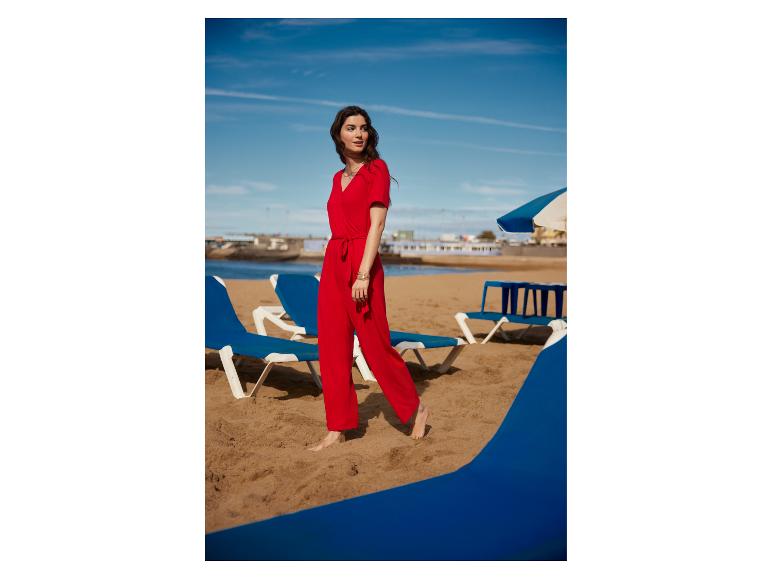 Vrouw in een rode jumpsuit loopt blootsvoets op een zandstrand met ligstoelen.