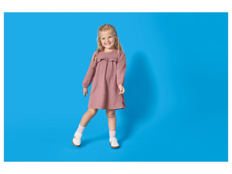 Lachend meisje in een roze jurk en witte sneakers op een blauwe achtergrond.