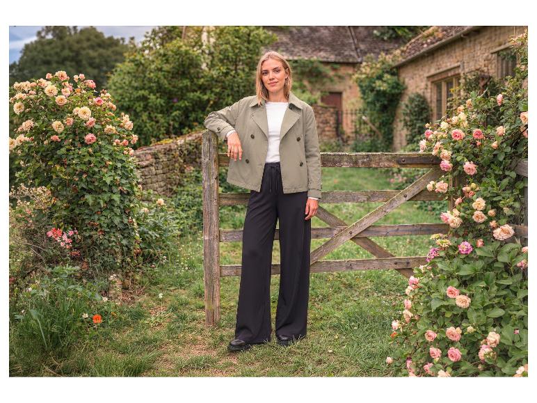 Vrouw in groene jas en zwarte wijde broek bij houten hek met rozen.