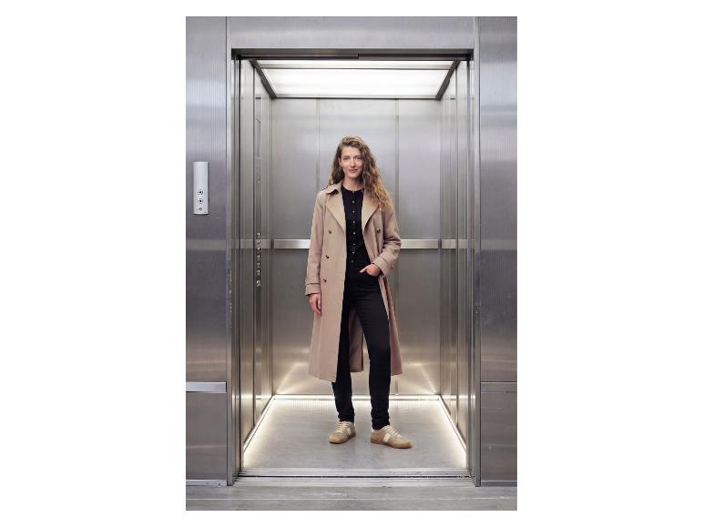 Vrouw in beige trenchcoat en zwarte broek in een lift.