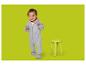 Lachende baby in grijze romper, staand naast een gele kruk.