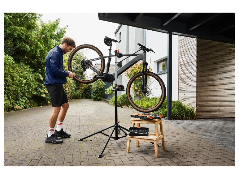 Man in blauw jack en zwarte korte broek repareert fiets op standaard met gereedschap op krukje.