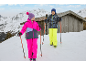 Twee kinderen in winterkleding met skistokken in een besneeuwd berglandschap met een houten hut.