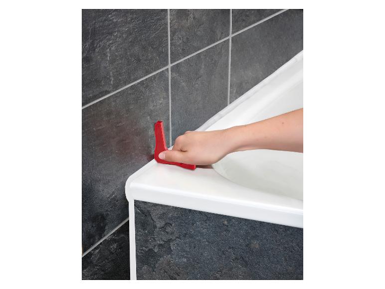 Een hand gebruikt een rood gereedschap om kit aan te brengen rond een wit bad.