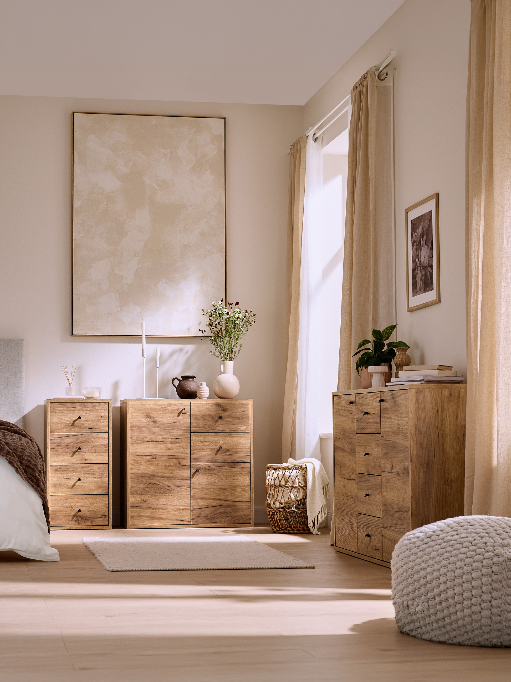Lichte slaapkamer met lichte houten ladekasten, bed en gebreide poef.