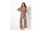 Vrouw in bruine jumpsuit met print, handen in de zij en leunend op een stoel.
