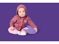 Baby in een roze vest met capuchon en roze broek, zittend op een paars kussen.