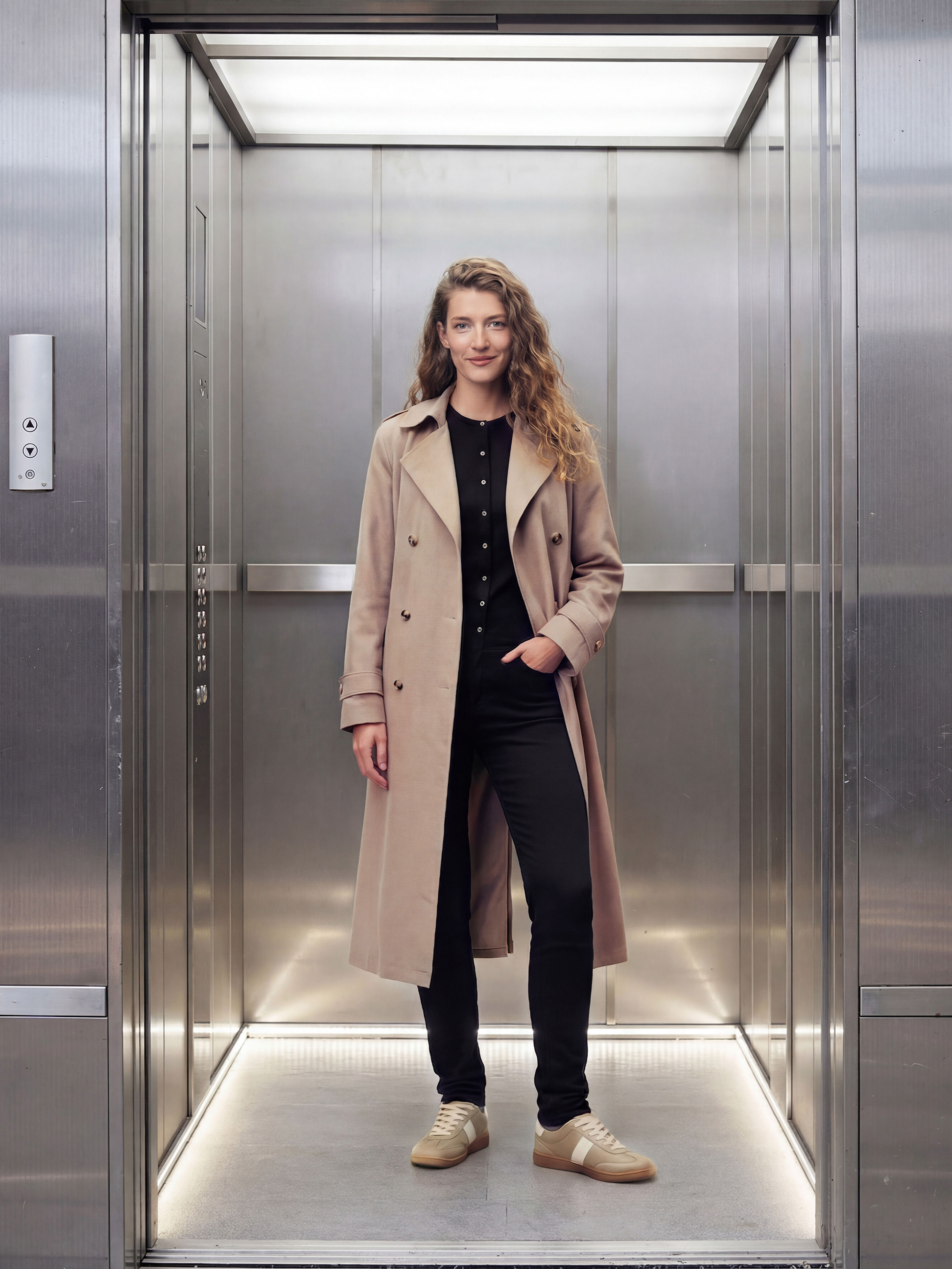 Vrouw in beige jas, zwarte blouse en broek in een lift.