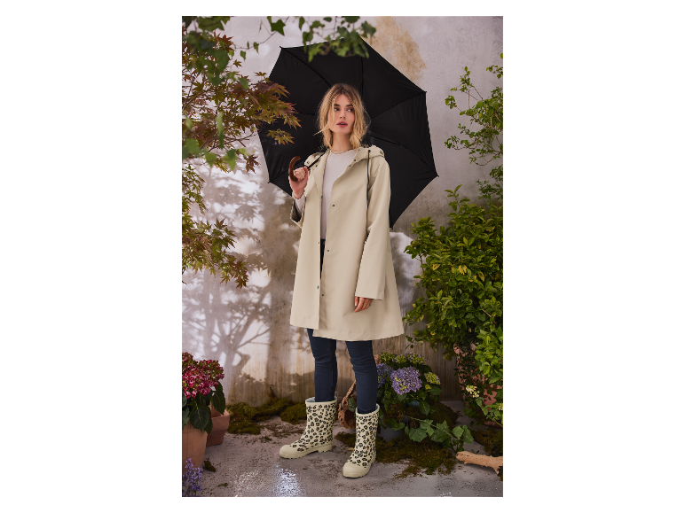 Vrouw in beige regenjas en laarzen met luipaardprint onder een zwarte paraplu.