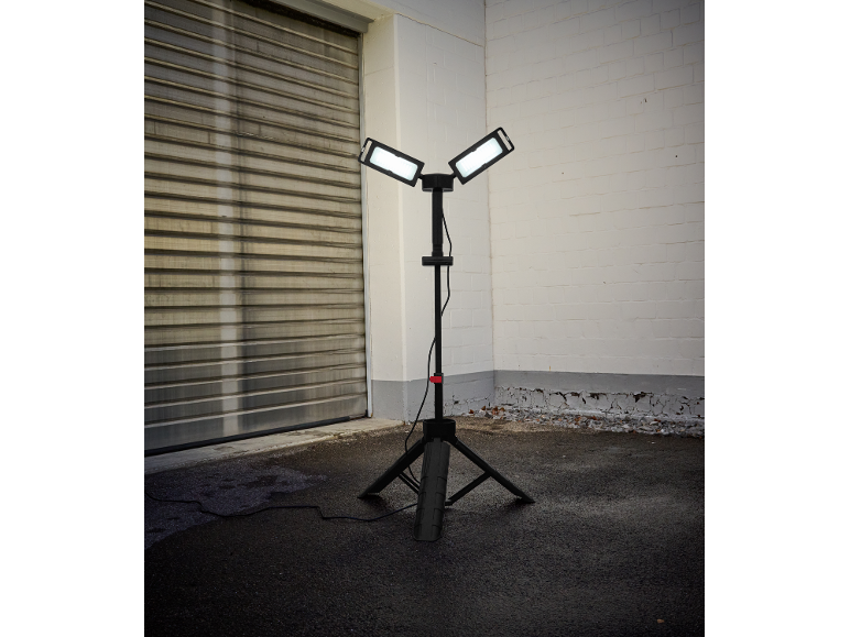 Statiefbouwlamp met twee verlichte panelen op asfalt naast een garagedeur en bakstenen muur.