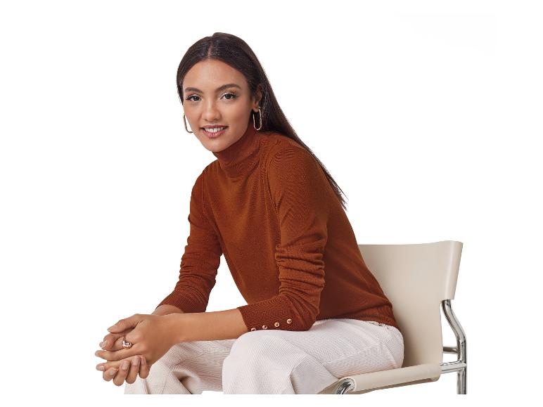 Een lachende vrouw draagt een bruine coltrui en een witte broek, zittend op een stoel.
