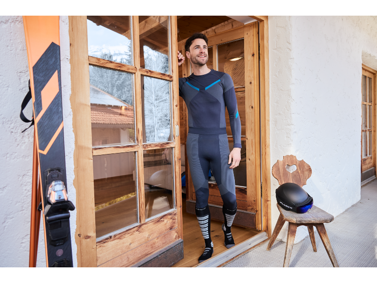 Man in thermokleding staat in deuropening, ski's en helm ernaast