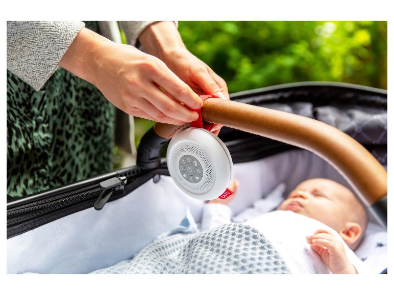 Ouder bevestigt een witte ZAZU geluidsmachine aan de kinderwagenbeugel boven een slapende baby.