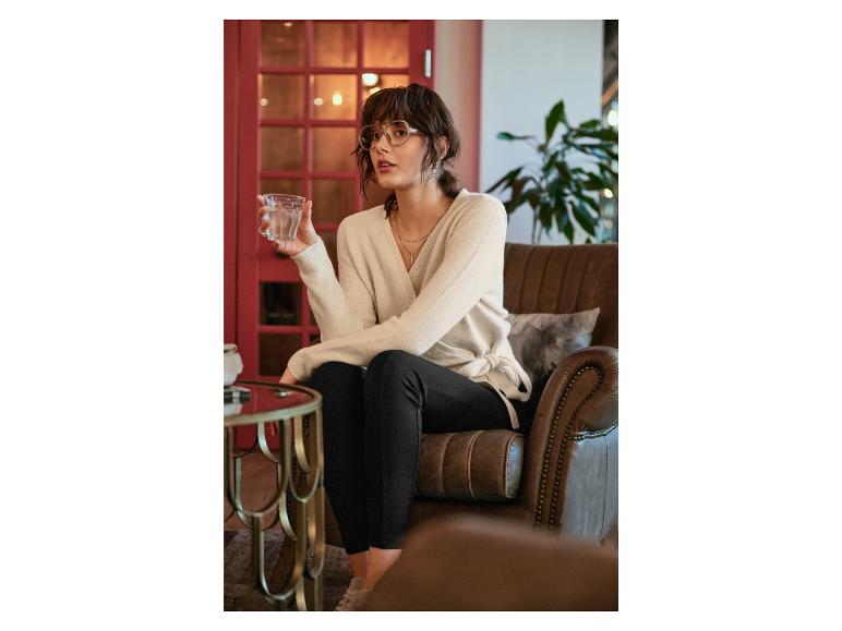 Een vrouw in een beige trui en zwarte broek zit op een leren fauteuil.