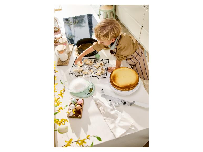 Kind versiert koekjes en een cheesecake op een aanrecht met bakgereedschap.