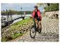 Vrouw fietst op een elektrische fiets langs de rivier, draagt een rode jas en helm.