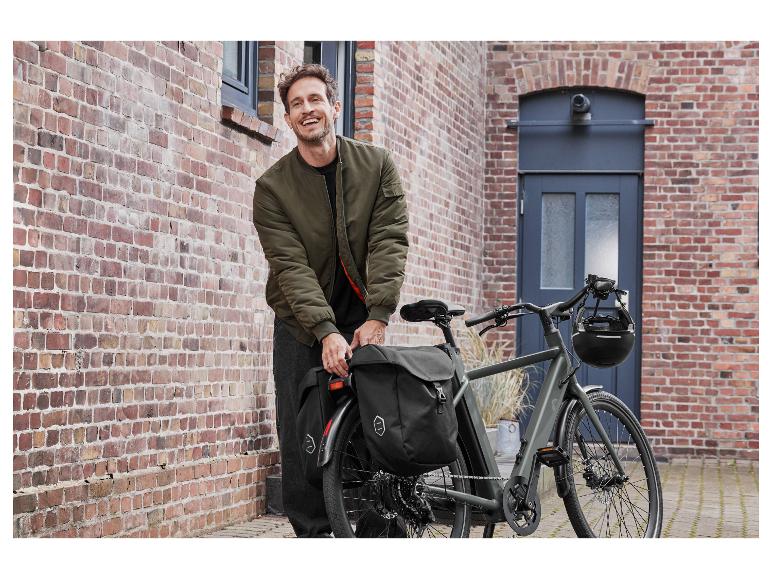 Een man staat naast een grijze elektrische fiets met twee tassen.