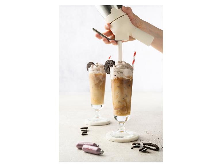 Hand spuit slagroom op ijskoffie met koekjes en gaspatronen op tafel.