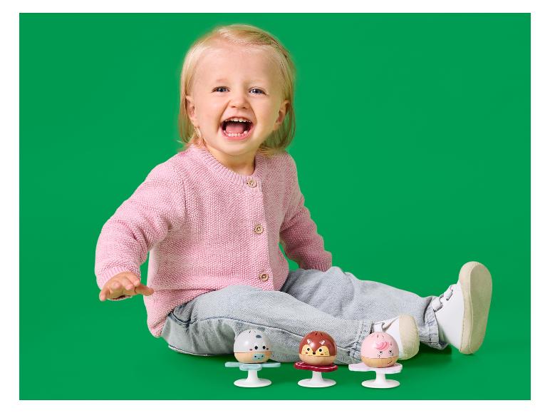 Blije baby in roze vestje en spijkerbroek, spelend met drie kleurrijke rammelaars.