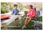 Campingtafel en stoelen, jongen leest boek, vrouw drinkt uit fles.