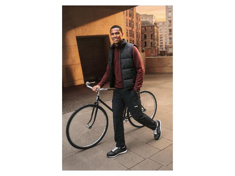 Man met fiets, draagt een bodywarmer, trui en cargobroek.
